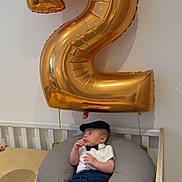 Ali participe au concours pour gagner de l'argent avec cette photo : baby, balloon, number_two, crib, pillow, bow_tie, cap, baby_clothes, indoor, child, white_wall, furniture, laying_down, cute, portrait, infant, celebration, gold, blue_pants, socks