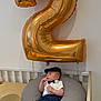 baby, balloon, number_two, crib, pillow, bow_tie, cap, baby_clothes, indoor, child, white_wall, furniture, laying_down, cute, portrait, infant, celebration, gold, blue_pants, socks