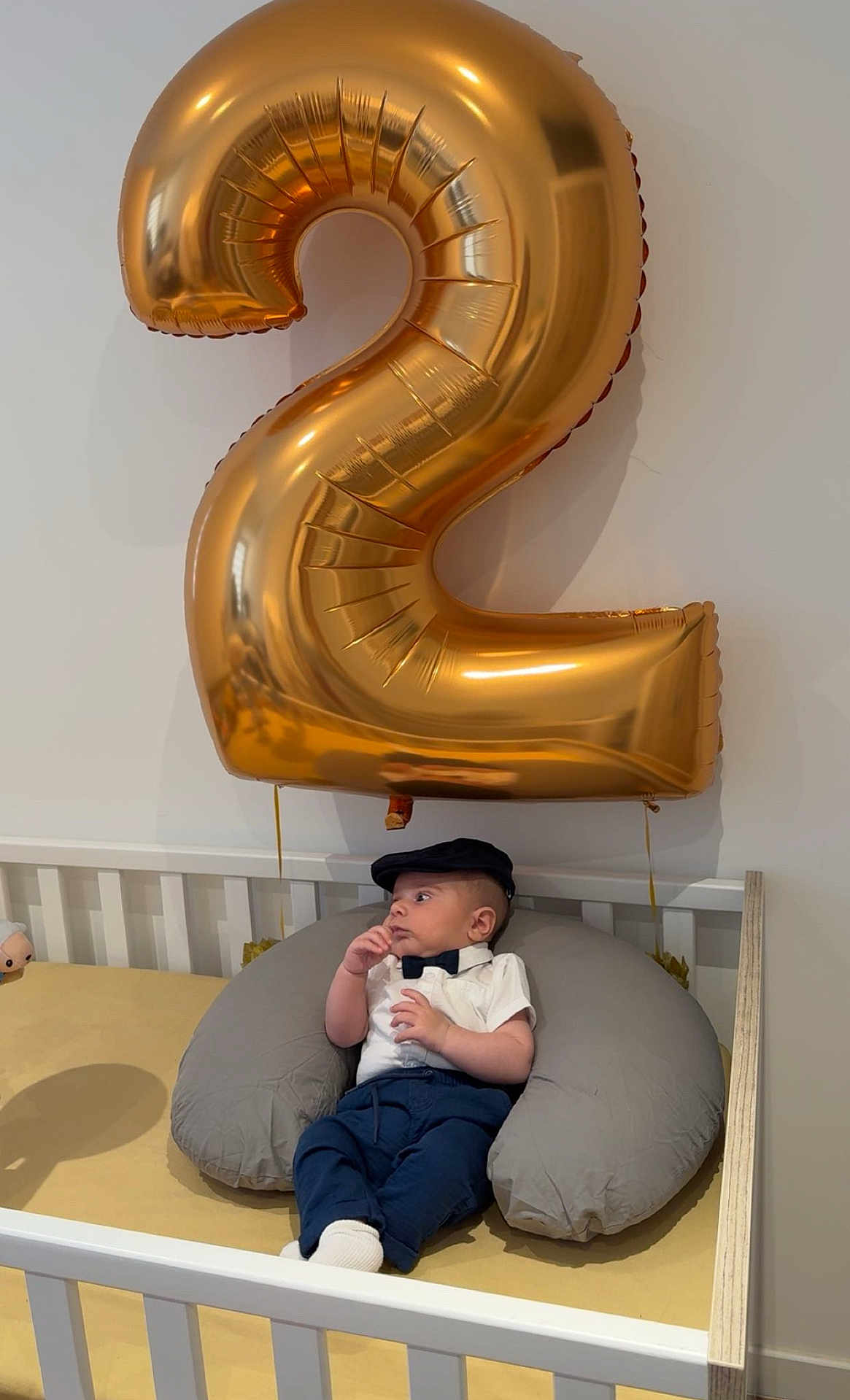 Ali participe au concours pour gagner de l'argent avec cette photo : baby, balloon, number_two, crib, pillow, bow_tie, cap, baby_clothes, indoor, child, white_wall, furniture, laying_down, cute, portrait, infant, celebration, gold, blue_pants, socks