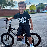 Mateo is registered to the contest to win money with this photo: child, boy, bicycle, bike, sneakers, black_tshirt, outdoor, street, smiling, shorts, daytime, suburban, tree, house, pavement, sky, clouds, person, young, happy