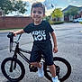 child, boy, bicycle, bike, sneakers, black_tshirt, outdoor, street, smiling, shorts, daytime, suburban, tree, house, pavement, sky, clouds, person, young, happy