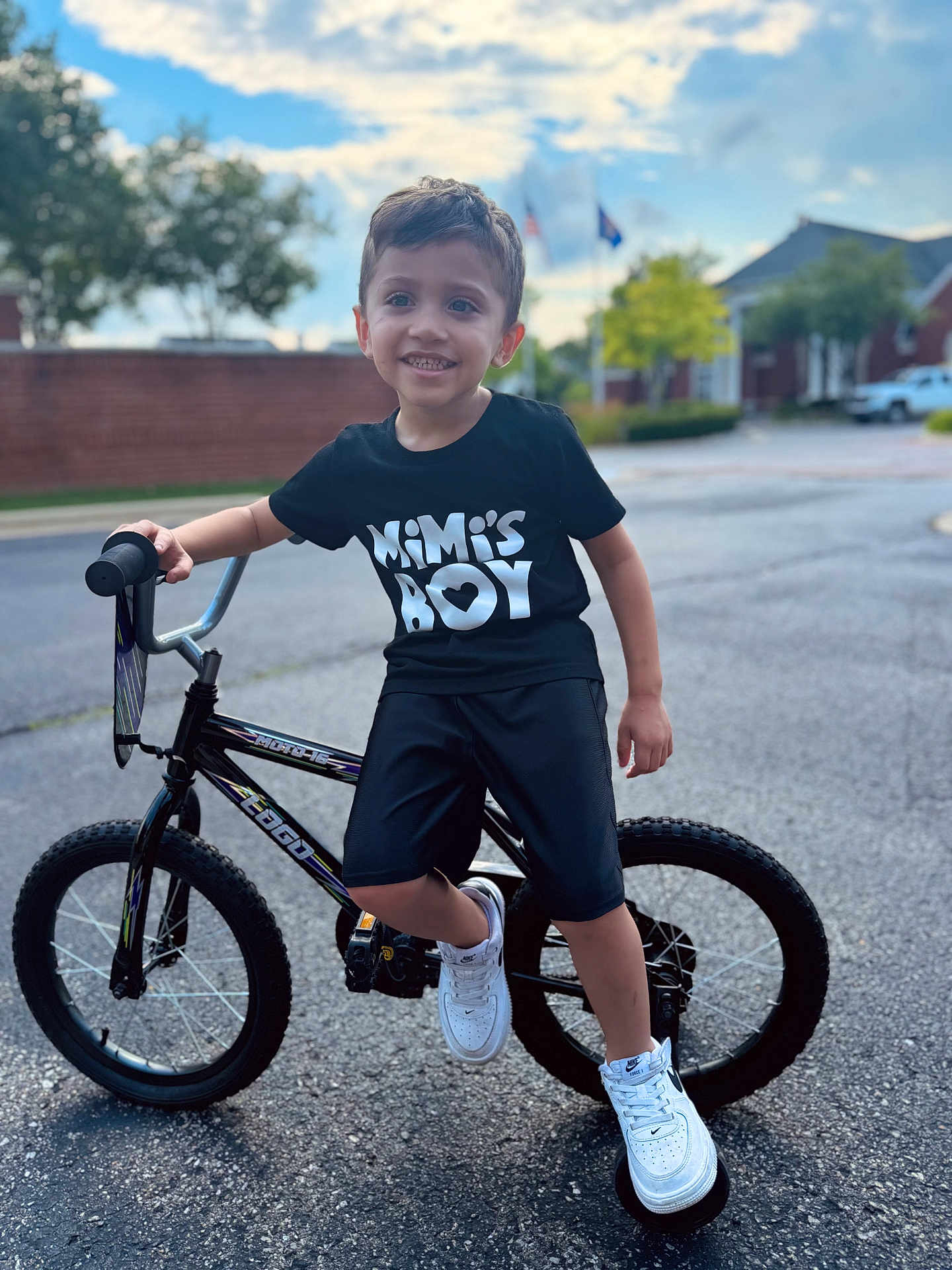 Mateo is registered to the contest to win money with this photo: child, boy, bicycle, bike, sneakers, black_tshirt, outdoor, street, smiling, shorts, daytime, suburban, tree, house, pavement, sky, clouds, person, young, happy