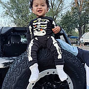 Dario joined the competition — help win amazing prizes! child, toddler, smiling, skeleton_pajamas, black_outfit, jeep, spare_tire, offroad_tire, outdoors, trees, camping, vehicle, socks, portrait, happy, park, young_child, curly_hair, playful, weekend