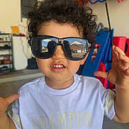 Dario joined the competition — help win amazing prizes! casual_clothing, champion_brand, child, colorful_background, curly_hair, face, floor, furniture, garage, hands_up, indoor, person, playful, portrait, reflection, smiling, sunglasses, toddler, toys, white_tshirt