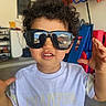 child, toddler, curly_hair, sunglasses, white_tshirt, champion_brand, indoor, garage, toys, playful, hands_up, smiling, person, casual_clothing, colorful_background, reflection, furniture, floor, face, portrait