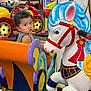 toddler, child, carousel, horse, amusement_ride, colorful, indoor, play_area, toy, face, curly_hair, expression, seat, entertainment, fun, kid, background, plastic, ride, cartoon_character