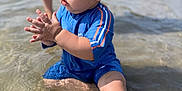 Dario is registered to the contest to win money with this photo: toddler, child, water, swimsuit, blue_clothing, curly_hair, beach, summer, sunny, sitting, shallow_water, clapping, outdoor, legs, person, nature, sand, play, fun, daytime