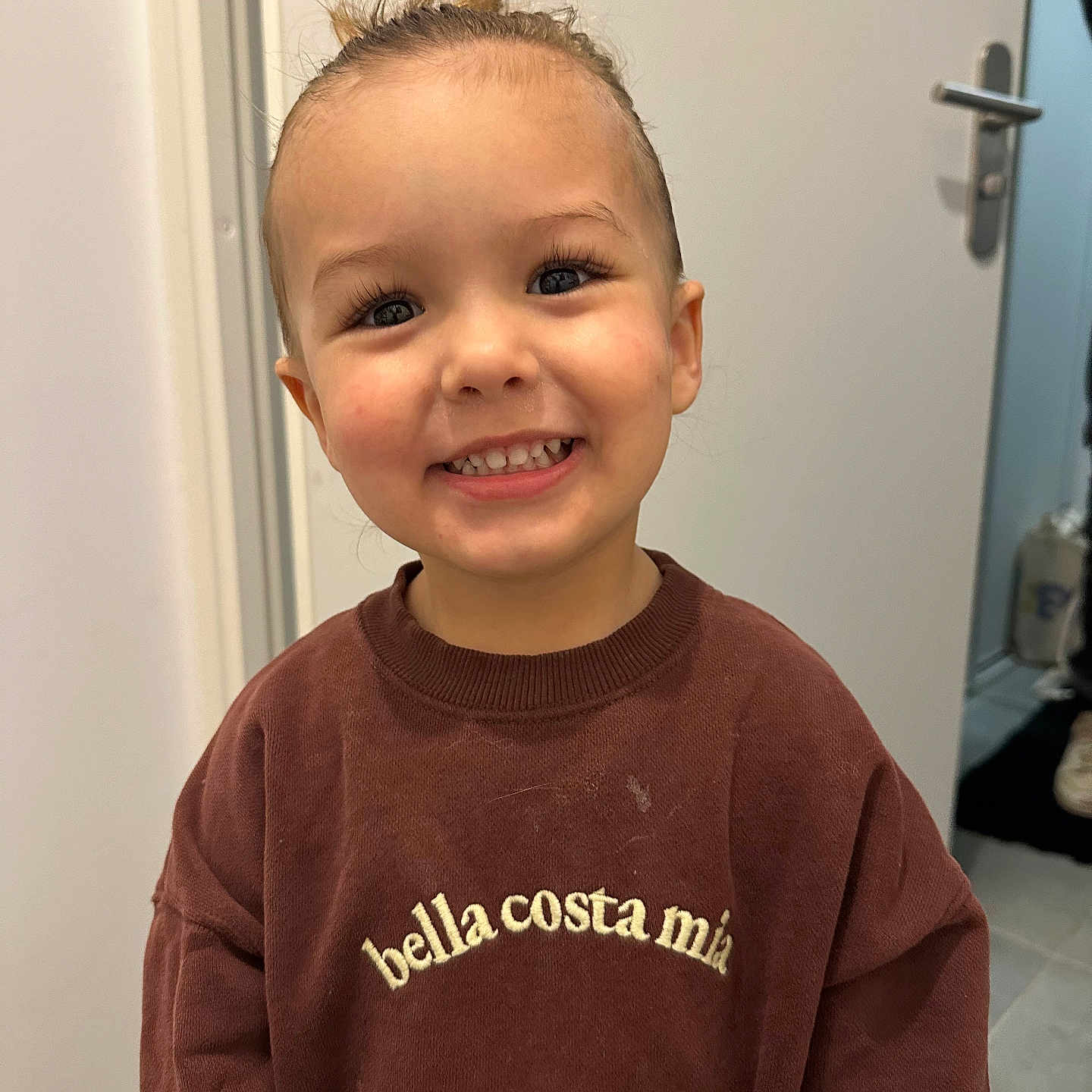 Mélyann a rejoint le concours — aidez-le/la à gagner de superbes lots ! child, smiling, indoor, sweatshirt, brown_clothing, hair_up, top_knot, face, happy, young, person, casual, door, floor, tile_floor, standing, portrait, cute, expression, human