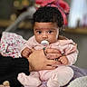 baby, infant, pacifier, curly_hair, hand, pink_clothing, sitting, indoor, soft_blanket, blurred_background, person, child, cute, portrait, cozy, adult_hand, furniture, warm, expression, comfort