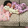 child, toddler, sleeping, pajamas, toy, stuffed_animal, couch, pillow, blanket, carpet, indoor, cozy, knitted_toy, purple, patterned, curly_hair, relaxing, home, comfort, nap