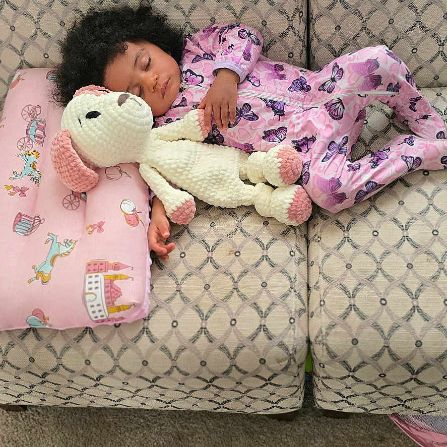 Neena joined the competition — help win amazing prizes! child, toddler, sleeping, pajamas, toy, stuffed_animal, couch, pillow, blanket, carpet, indoor, cozy, knitted_toy, purple, patterned, curly_hair, relaxing, home, comfort, nap