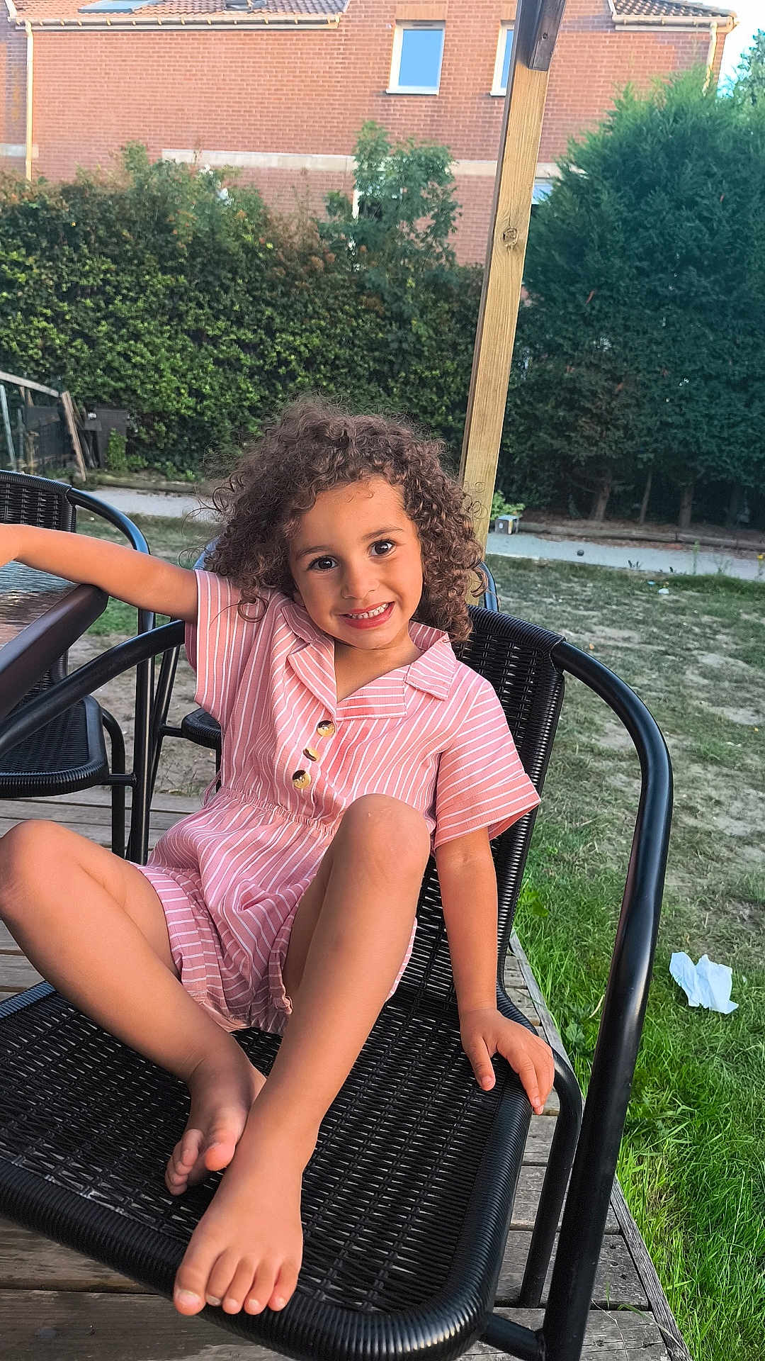 Siham participe au concours pour gagner de l'argent avec cette photo : child, smiling, curly_hair, pink_outfit, striped_clothing, black_chair, outdoor, garden, grass, hedge, brick_house, wooden_deck, relaxed, barefoot, happy, summer, daylight, portrait, casual, nature