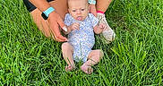Leilani is registered to the contest to win money with this photo: baby, adult, grass, outdoor, tattoo, watch, onesie, person, child, greenery, nature, casual, supporting, legs, hands, sitting, footwear, shorts, tank_top, skin