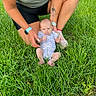 baby, adult, grass, outdoor, tattoo, watch, onesie, person, child, greenery, nature, casual, supporting, legs, hands, sitting, footwear, shorts, tank_top, skin