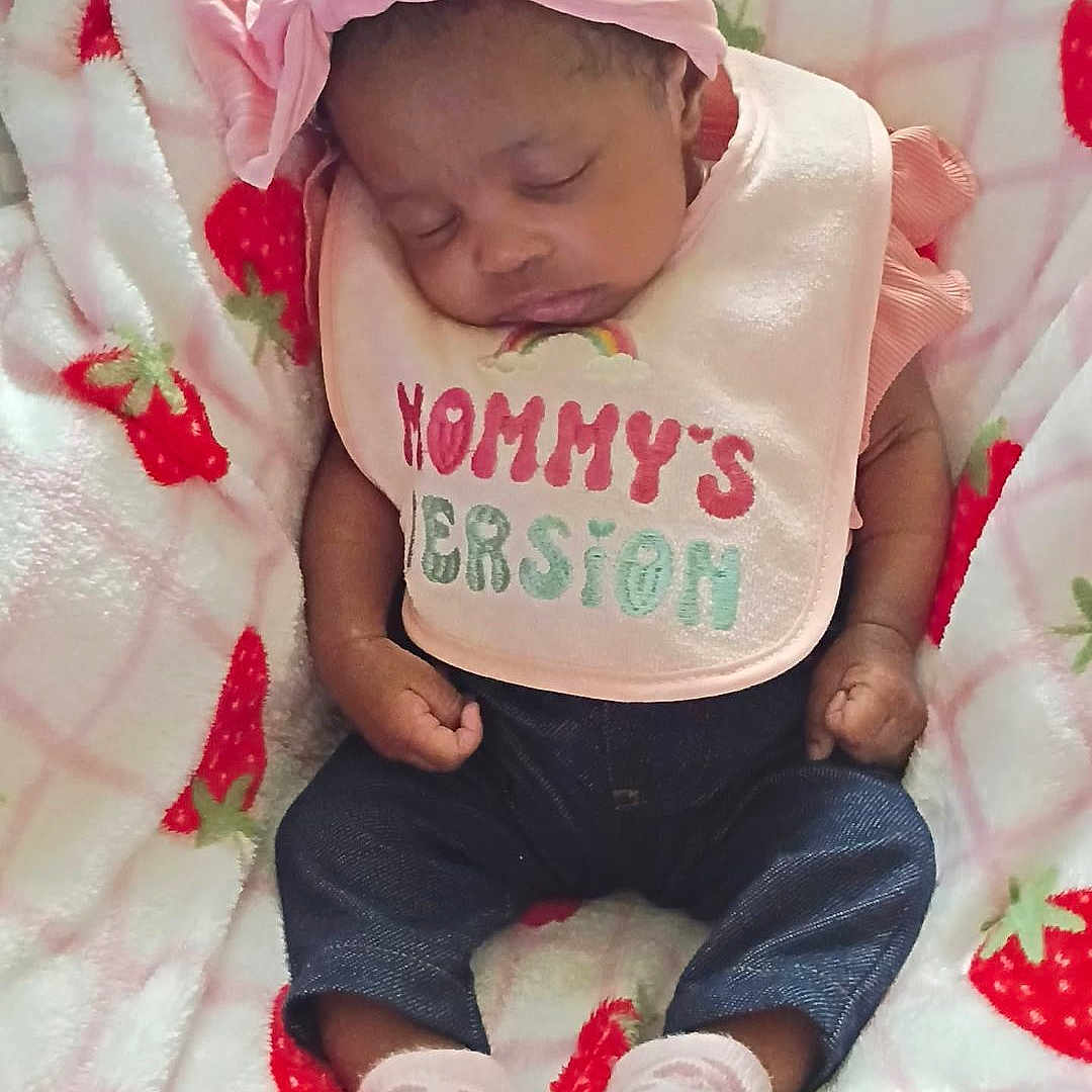 Anijah is registered to the contest to win money with this photo: baby, newborn, sleeping, portrait, pink, headband, bow, bib, clothing, blanket, strawberry, infant, face, closed_eyes, indoors, cozy, lying_down, socks, dark_skin, family