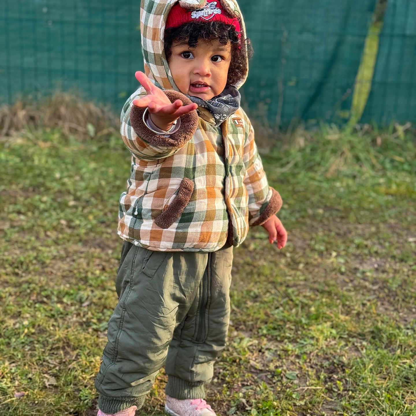 Liria a rejoint le concours — aidez-le/la à gagner de superbes lots ! beanie, casual_clothing, child, cold_weather, curly_hair, expression, fence, gesture, grass, green_pants, hat, hood, jacket, nature, outdoor, playful, portrait, sneakers, standing, toddler