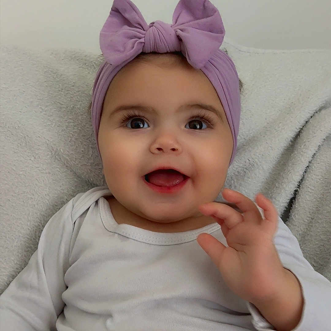 Tylia participe au concours pour gagner de l'argent avec cette photo : baby, blanket, child, clothing, cute, eyes, face, hand, happy, headband, indoor, infant, onesie, portrait, purple_bow, sitting, skin, smiling, soft, waving