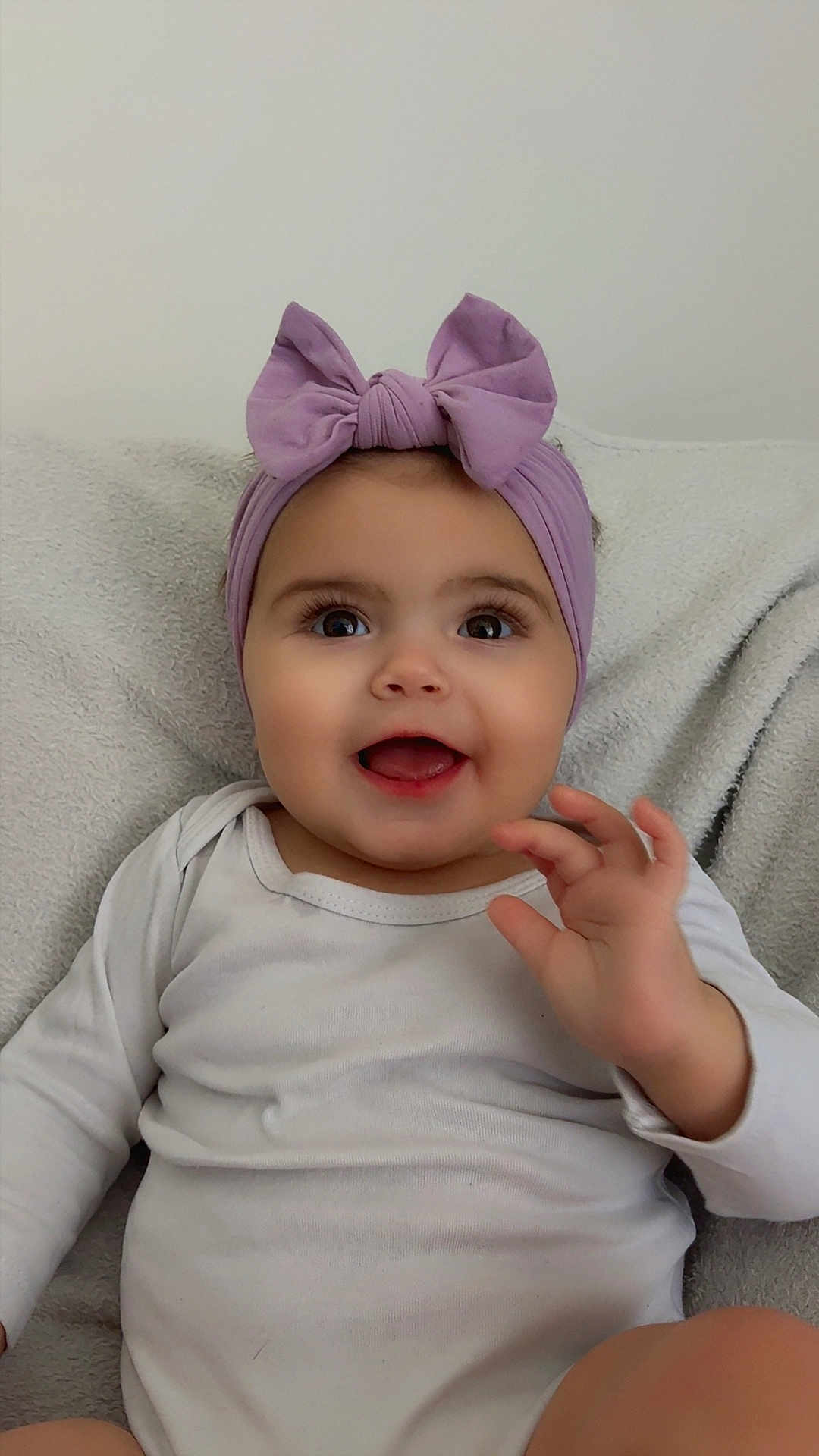 Tylia participe au concours pour gagner de l'argent avec cette photo : baby, child, smiling, headband, purple_bow, onesie, happy, cute, infant, waving, hand, face, eyes, skin, clothing, sitting, blanket, soft, indoor, portrait