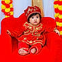 Nidhyanti joined the competition — help win amazing prizes! baby, child, red_clothing, traditional_outfit, hat, plush_chair, flower_garland, orange, yellow, red, decor, portrait, cute, indoor, festive, sitting, face, expression, curious, young_child