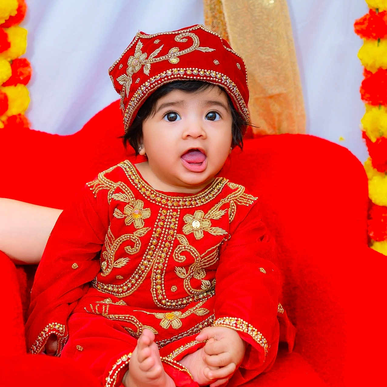 Nidhyanti is registered to the contest to win money with this photo: baby, bright_colors, celebration, chair, child, cute, decorations, expression, face, foot, hand, hat, indoors, orange_flowers, portrait, red_clothing, sitting, traditional_outfit, yellow_flowers, young_child