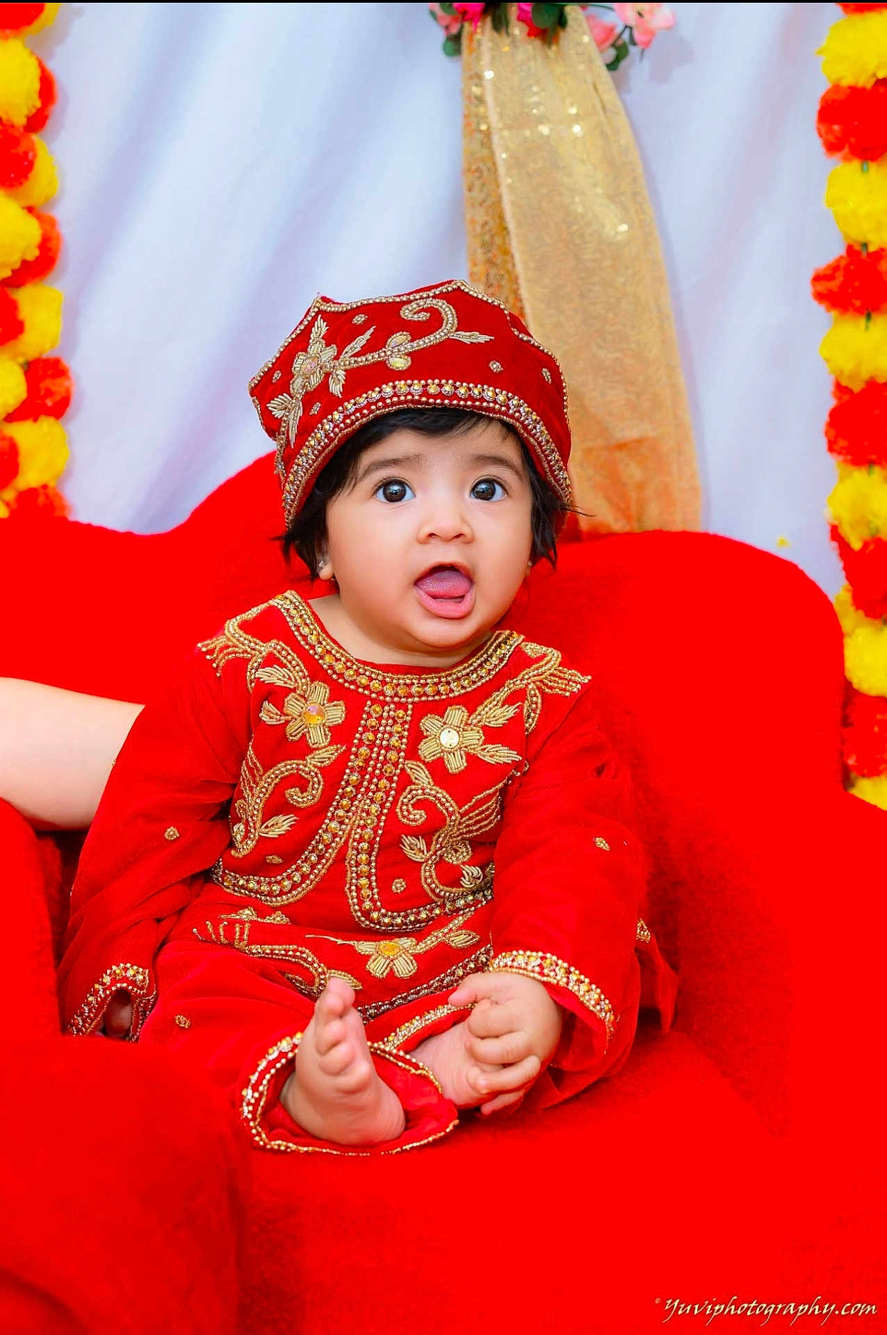 Nidhyanti is registered to the contest to win money with this photo: baby, child, red_clothing, traditional_outfit, hat, sitting, chair, face, expression, decorations, yellow_flowers, orange_flowers, portrait, indoors, cute, foot, hand, bright_colors, celebration, young_child