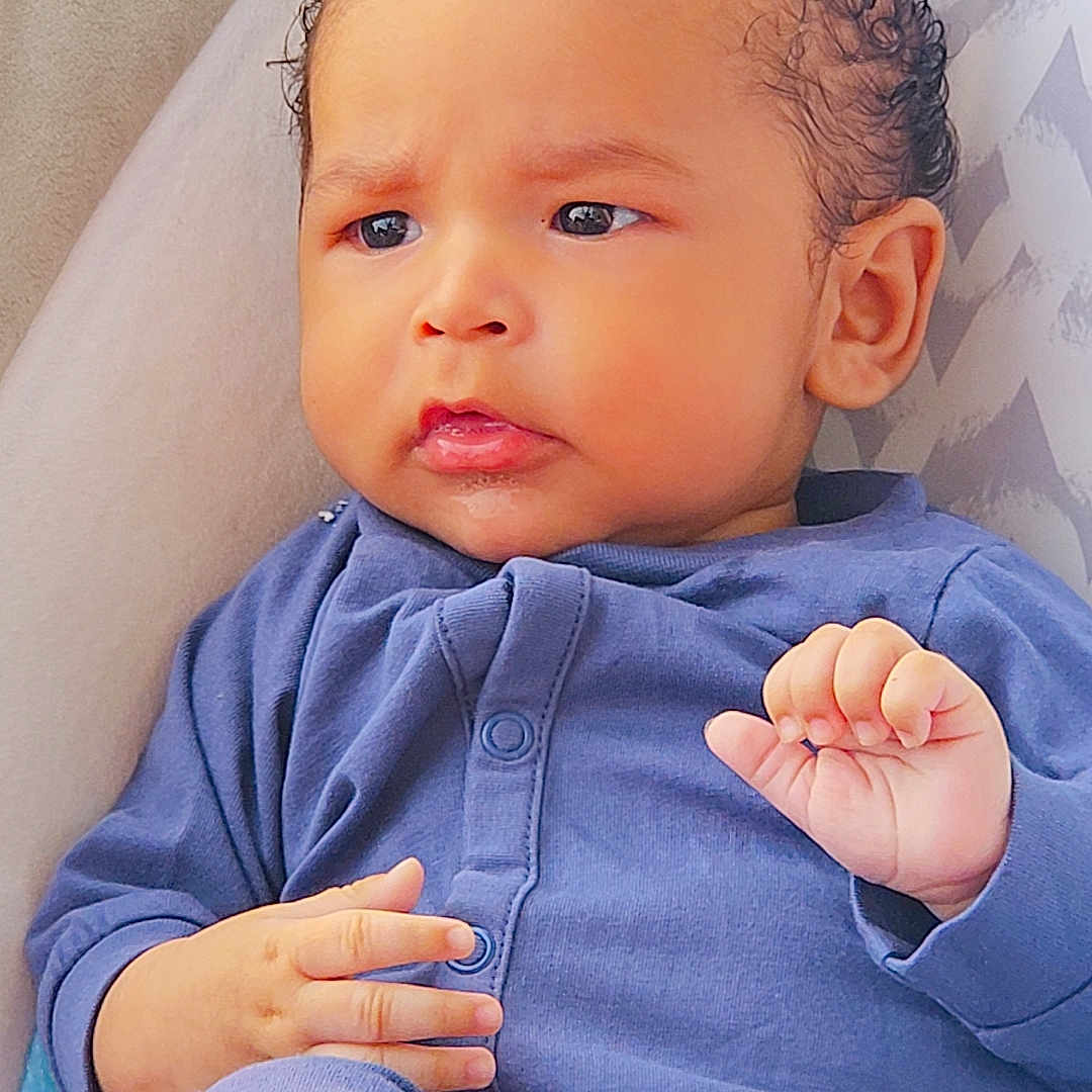 Ezmari is registered to the contest to win money with this photo: baby, blue_clothing, child, closeup, comfortable, cozy, curly_hair, cushion, cute, expression, face, hand, indoors, infant, pajamas, patterned_background, portrait, seated, skin, young_child