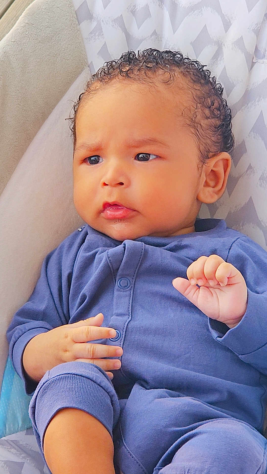 Ezmari is registered to the contest to win money with this photo: baby, infant, child, pajamas, blue_clothing, curly_hair, indoors, cushion, patterned_background, seated, hand, face, expression, skin, portrait, young_child, comfortable, cozy, closeup, cute