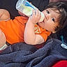 baby, child, infant, bottle, feeding, orange_shirt, blanket, red_blanket, couch, lying_down, portrait, face, eyes, hair, smiling, indoors, cozy, casual, 家庭, photograph
