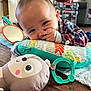 Baby Jose is registered to the contest to win money with this photo: baby, child, infant, pillow, toy, plush_toy, mirror, floor, hand, face, smiling, indoors, clothing, plaid_shirt, wooden_floor, cute, playtime, home, person, colorful