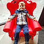 boy, casual_clothing, child, floor, furniture, happy, indoor, interior, jacket, natural_light, pants, person, portrait, red_chair, relaxed, seat, smiling, sneakers, window, young