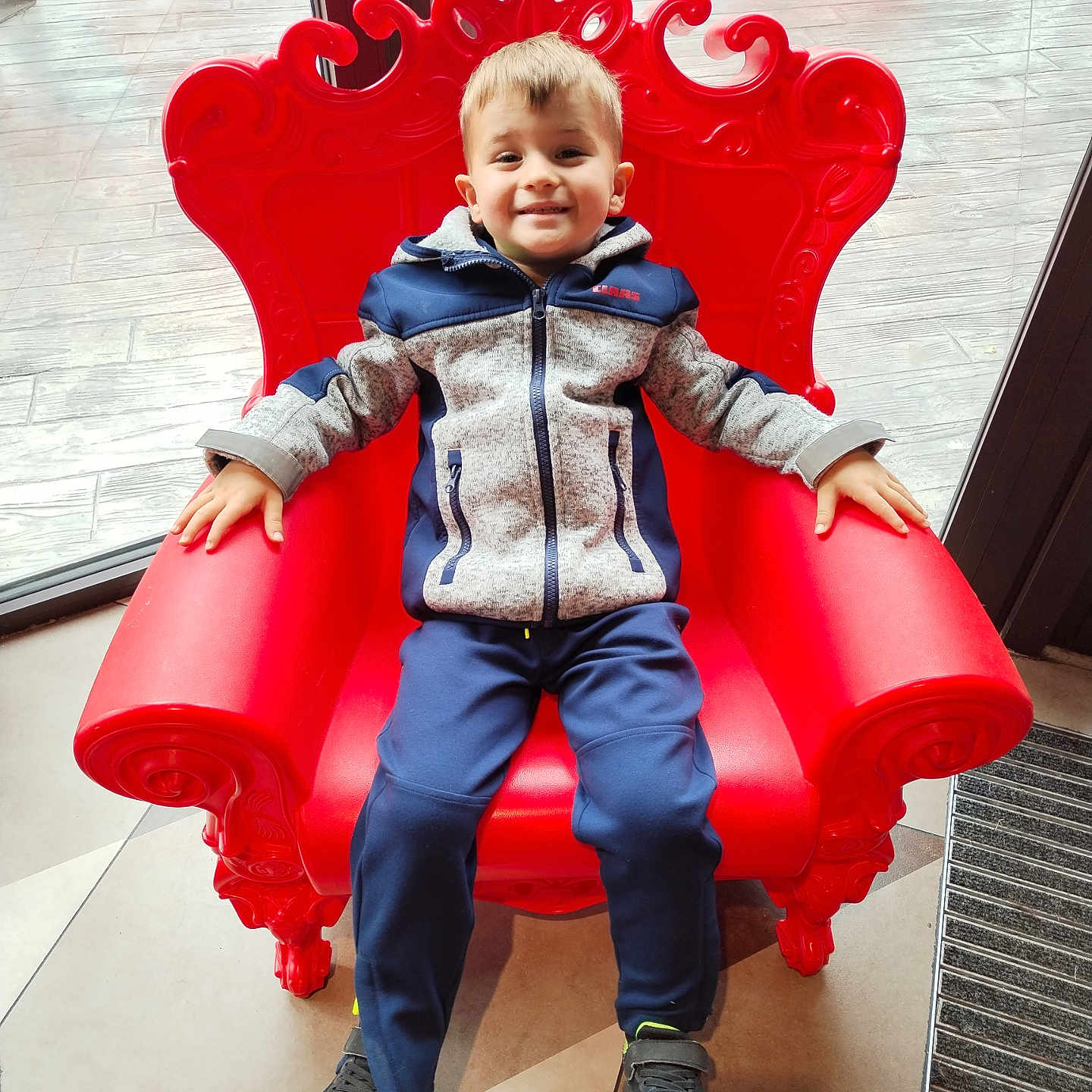Naël participe au concours pour gagner de l'argent avec cette photo : boy, casual_clothing, child, floor, furniture, happy, indoor, interior, jacket, natural_light, pants, person, portrait, red_chair, relaxed, seat, smiling, sneakers, window, young
