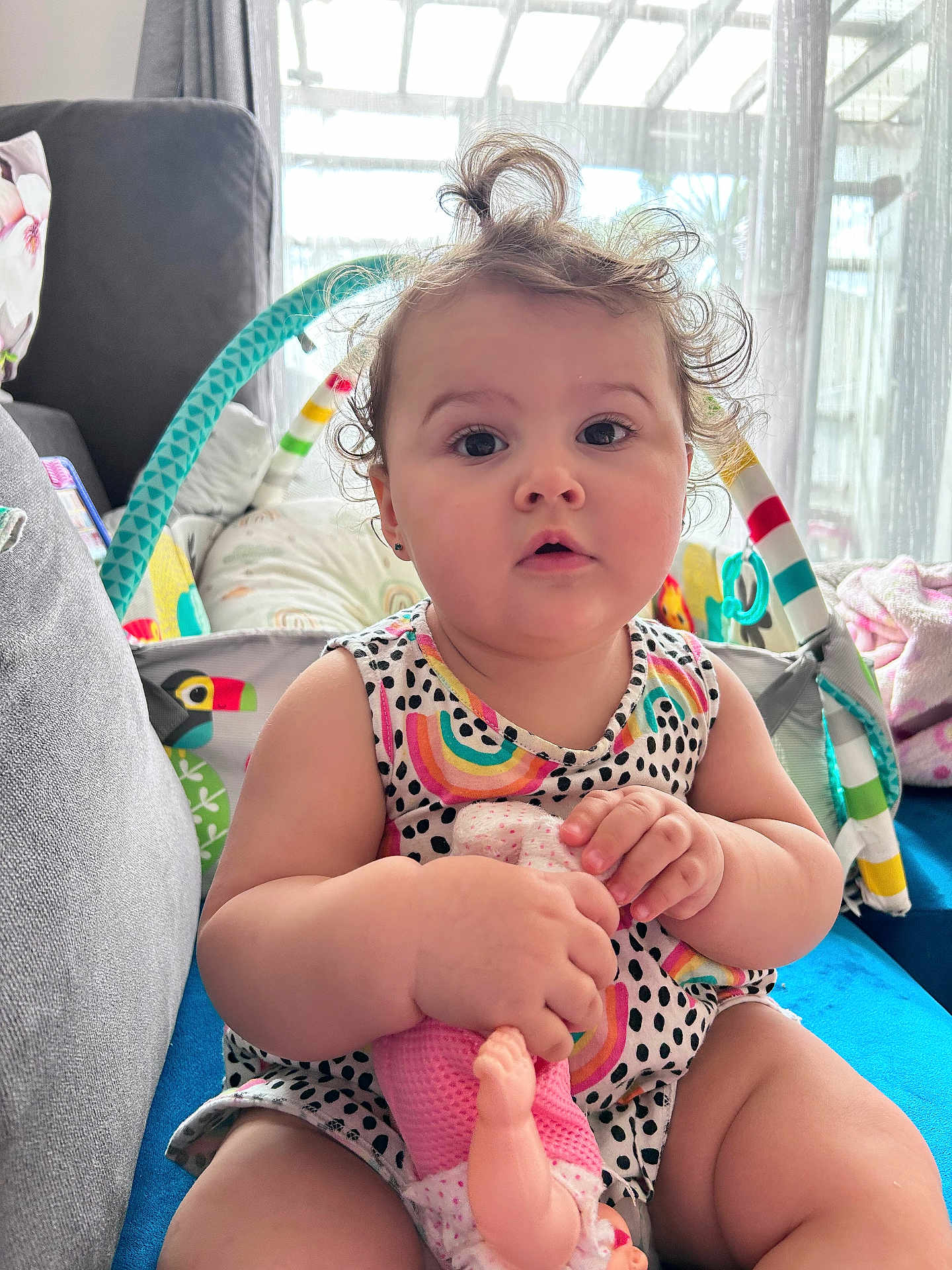 Sarah is registered to the contest to win money with this photo: baby, child, doll, curly_hair, sitting, indoor, couch, toy, colorful, cute, infant, clothing, patterned_dress, face, hands, curtains, window, soft_light, home, person