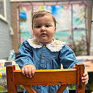 Sarah is registered to the contest to win money with this photo: toddler, child, denim_dress, wooden_chair, embroidery, curly_hair, portrait, indoor, hand, face, expression, background_blur, colorful_mural, buttoned_dress, young_child, holding, person, cute, casual_clothing, curious