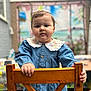 toddler, child, denim_dress, wooden_chair, embroidery, curly_hair, portrait, indoor, hand, face, expression, background_blur, colorful_mural, buttoned_dress, young_child, holding, person, cute, casual_clothing, curious