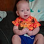 Tyberius is registered to the contest to win money with this photo: baby, child, infant, sitting, clothing, orange_shirt, blue_shorts, blanket, couch, indoor, face, hands, legs, footwear, wooden_wall, fabric, comfort, casual, young, cute