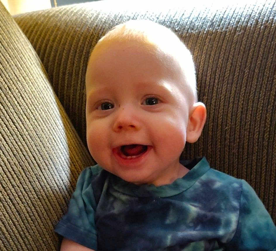 Tyberius is registered to the contest to win money with this photo: baby, child, smiling, face, eyes, mouth, shirt, tie_dye, couch, furniture, indoors, happy, young, person, portrait, sitting, cute, skin, head, expression
