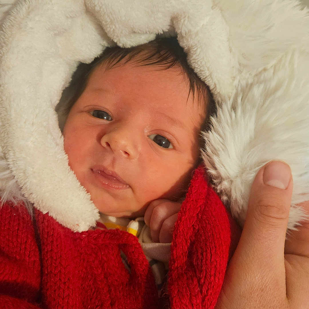 Caspian is registered to the contest to win money with this photo: baby, closeup, cute, expression, eyes, face, hand, headgear, holding, indoor, infant, knitwear, newborn, person, portrait, red_sweater, skin, soft_texture, warm_clothing, white_hood