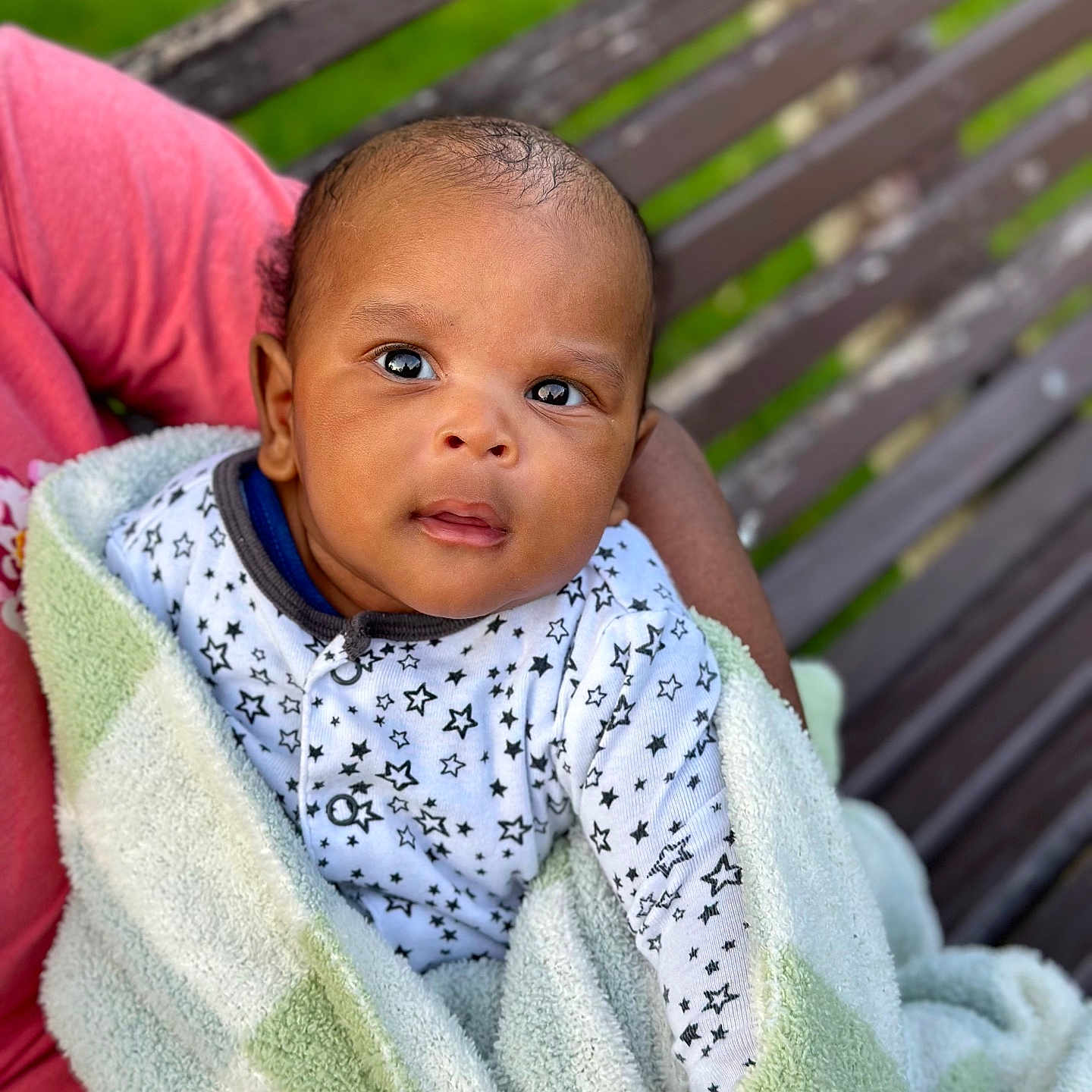 Yann is registered to the contest to win money with this photo: baby, bench, blanket, closeup, clothing, curious, cute, expression, face, grass, green, hand, infant, outdoor, person, portrait, soft_texture, wood, wrapped, young_child