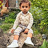 child, toddler, portrait, sitting, outdoors, garden, tree, tree_trunk, greenery, plants, leaves, clothing, shirt, suspenders, plaid_pants, sneakers, socks, direct_gaze, natural_light, garden_pots