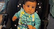 Alen is registered to the contest to win money with this photo: baby, child, infant, stroller, portrait, sitting, indoors, home, plaid, overalls, colorful_clothing, star_pattern, wide_eyed, looking_at_camera, brown_hair, cozy, houseplant, curtains, furniture, photography