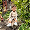 child, toddler, portrait, outdoor, garden, tree_stump, sitting, formal_clothing, button_up_shirt, shorts, suspenders, sneakers, socks, curly_hair, serious_expression, greenery, plants, rustic_shed, flower_pot, natural_light