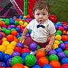 toddler, child, ball_pit, colorful_balls, indoor, play_area, white_shirt, bow_tie, maroon_shorts, plastic_balls, curious, sitting, playful, young_child, person, fun, entertainment, cute, expression, recreation