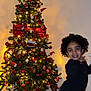 child, christmas_tree, holiday_decor, ornaments, lights, red_bow, indoor, smiling, posing, jeans, black_sweater, socks, wooden_floor, festive, celebration, smile, young_child, curly_hair, home, happy