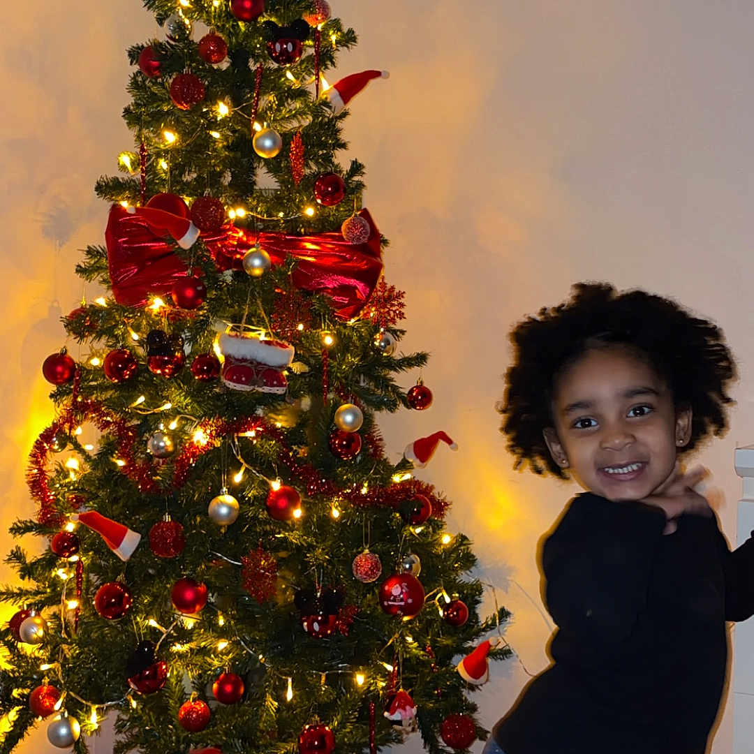 Neilah a rejoint le concours — aidez-le/la à gagner de superbes lots ! black_sweater, celebration, child, christmas_tree, curly_hair, festive, happy, holiday_decor, home, indoor, jeans, lights, ornaments, posing, red_bow, smile, smiling, socks, wooden_floor, young_child