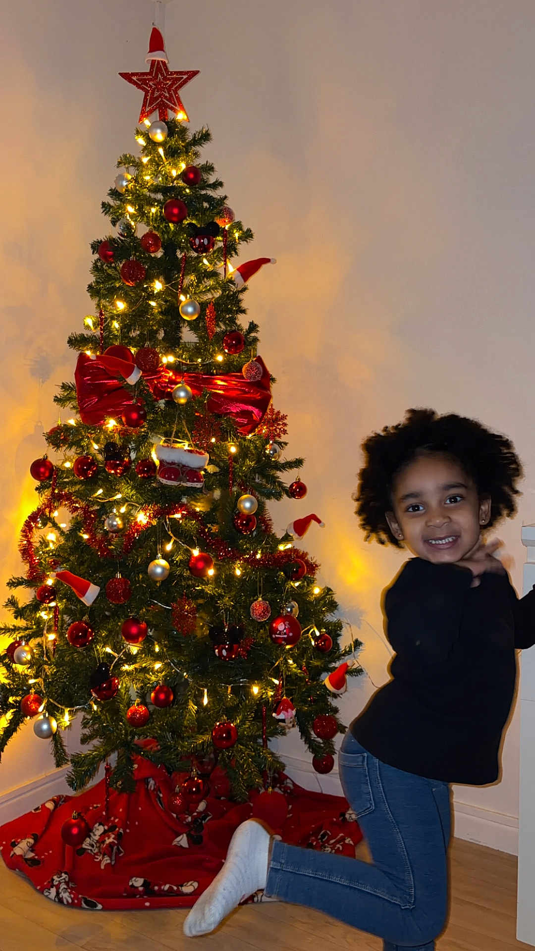 Neilah a rejoint le concours — aidez-le/la à gagner de superbes lots ! child, christmas_tree, holiday_decor, ornaments, lights, red_bow, indoor, smiling, posing, jeans, black_sweater, socks, wooden_floor, festive, celebration, smile, young_child, curly_hair, home, happy