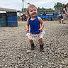 toddler, child, blue_top, lace_skirt, cowboy_boots, gravel, outdoor, event, people, folding_chairs, smile, pony_tail, casual, daytime, happy, standing, ground, sky, fence, hat