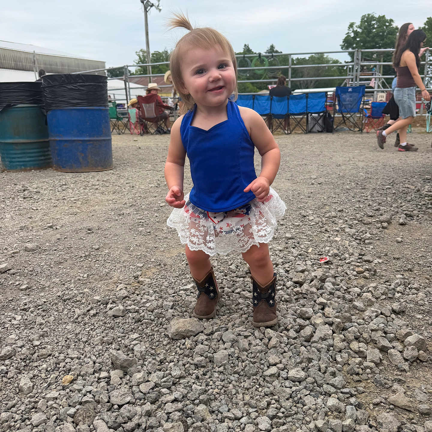 Alexandria Gabrielle is registered to the contest to win money with this photo: toddler, child, blue_top, lace_skirt, cowboy_boots, gravel, outdoor, event, people, folding_chairs, smile, pony_tail, casual, daytime, happy, standing, ground, sky, fence, hat