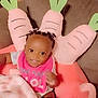 baby, child, smiling, braids, bib, blanket, couch, pillow, carrot_pillow, indoors, cute, happy, seated, infant, clothing, home, soft_toy, person, portrait, cozy
