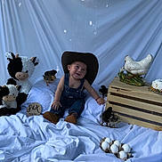 Juanluis is registered to the contest to win money with this photo: baby, cowboy_hat, denim_overalls, plush_cow, chicks, wooden_crate, ceramic_hen, eggs, blanket, smiling, sitting, cute, indoors, decor, child, toy, animal, farm_theme, portrait, happy