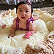 Selene is registered to the contest to win money with this photo: baby, child, infant, purple_clothing, pink_bows, soft_cushion, carpet, bed, indoor, cute, holding_object, pink_object, portrait, face, hands, wide_eyes, open_mouth, skin, short_hair, person